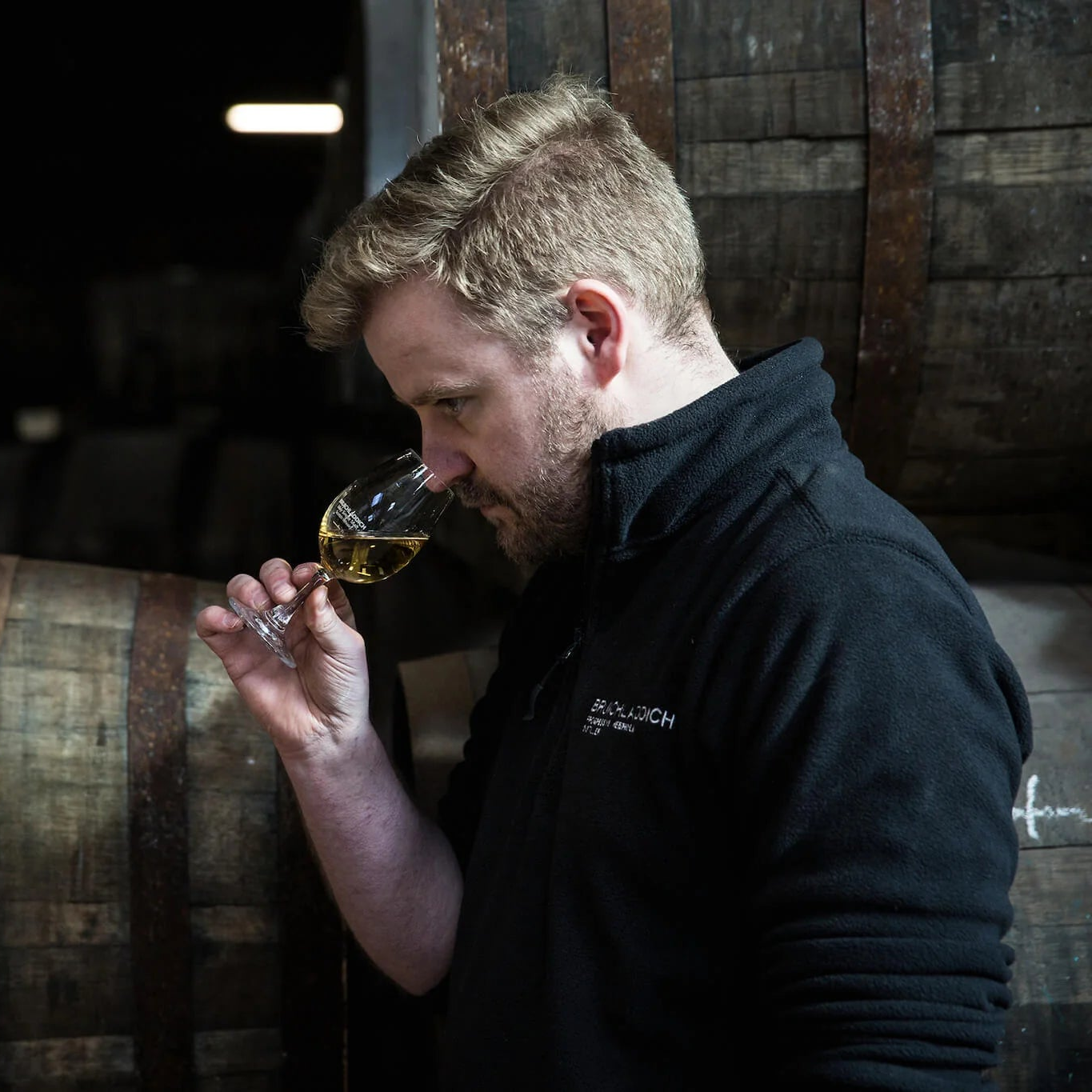 Adam Hannett, Head Distiller at Bruichladdich Distillery, nosing a Copita glass containing whisky in a warehouse surrounded by casks.