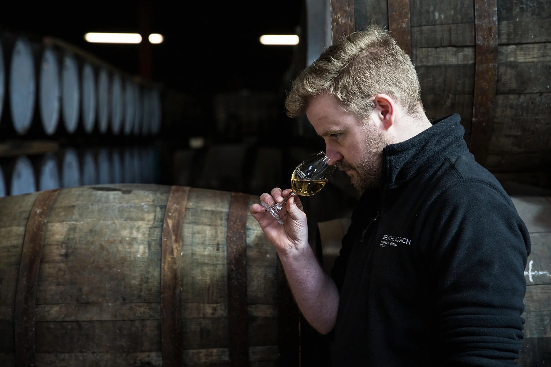 Adam Hannett, Head Distiller at Bruichladdich Distillery, nosing a Copita glass containing whisky in a warehouse surrounded by casks.